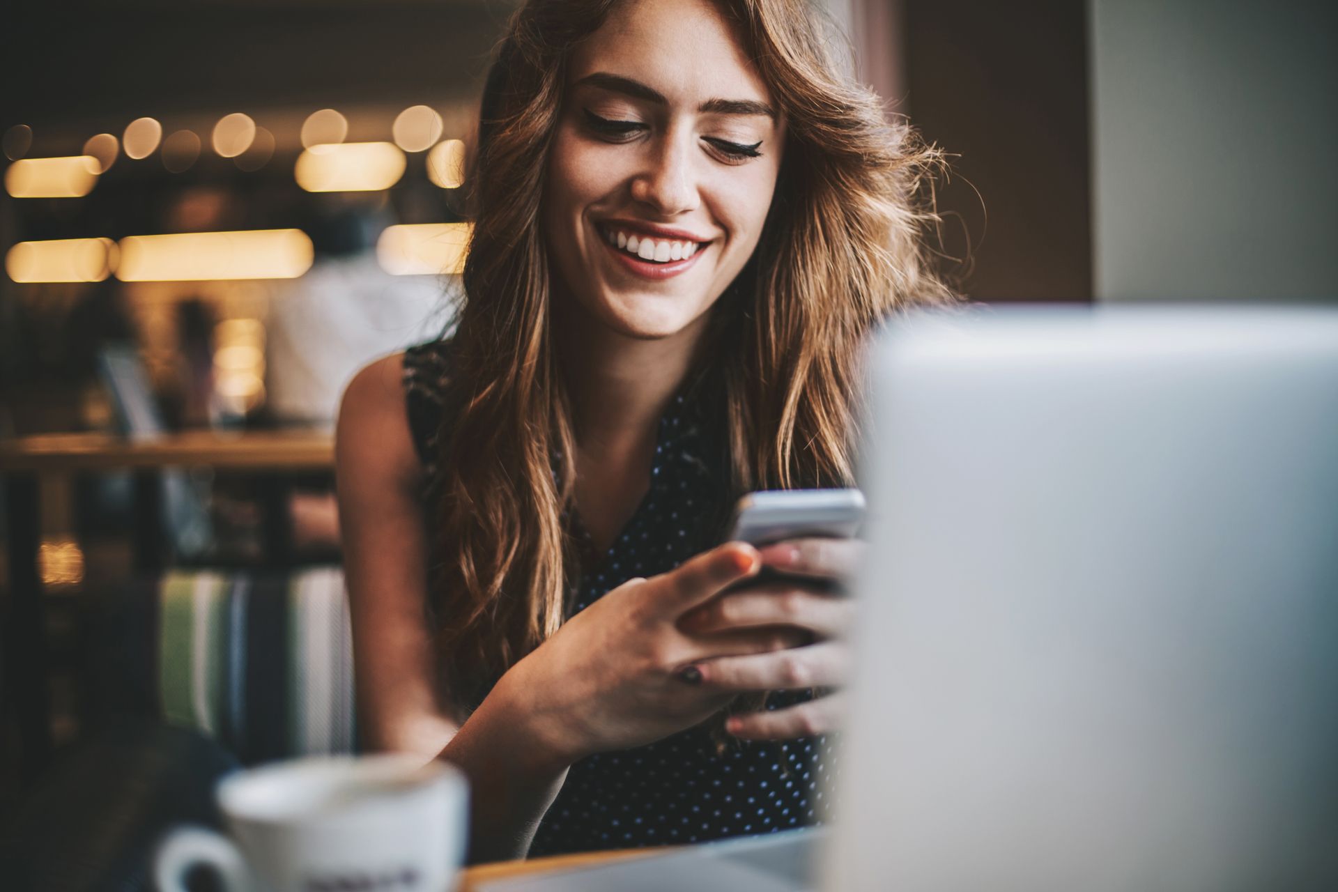 Woman smiling while looking at her smartphone in a modern home office setup with laptop and coffee cup.